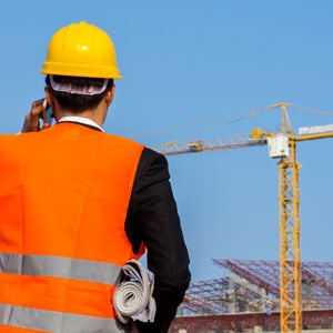A man in an orange vest and hard hat talking on a cell phone, possibly discussing work or receiving important instructions. - Leep Tescher Helfman and Zanze